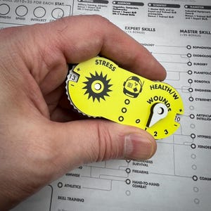 May include: A bright yellow game tracker with black text and graphics. It features a gear-shaped dial labelled STRESS with a plus and minus sign, a central astronaut icon, and a HEALTH/WOUNDS dial with numbers 1 through 10 and a plus and minus sign.