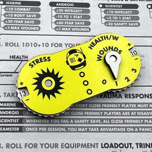 Puede incluir: Un rastreador de juego amarillo de forma ovalada con texto y gr&aacute;ficos en negro. Cuenta con un dial de estr&eacute;s con borde dentado, un contador de salud y heridas con un puntero met&aacute;lico y un icono de astronauta central.