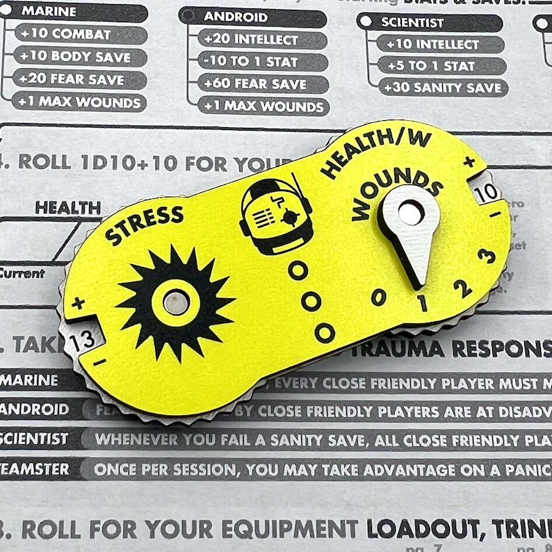 Puede incluir: Un rastreador de juego amarillo de forma ovalada con texto y gr&aacute;ficos en negro. Cuenta con un dial de estr&eacute;s con borde dentado, un contador de salud y heridas con un puntero met&aacute;lico y un icono de astronauta central.