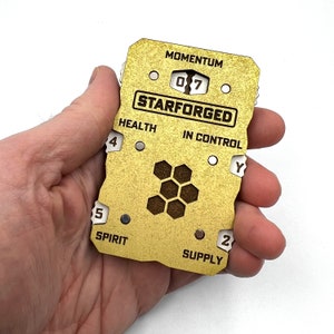 May include: A gold-coloured wooden player tracker for the board game Starforged. The tracker has a honeycomb design in the centre and includes spaces for health, spirit, supply, momentum, and in control.