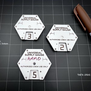 May include: Three hexagonal metal Universal Supply Gauges with the text "UNIVERSAL SUPPLY GAUGE" and "AUTHORIZED CREW USE ONLY". The gauges display the numbers 3, 2, and 5. One gauge has "Ammo" written on it. A brown marker is in the background.