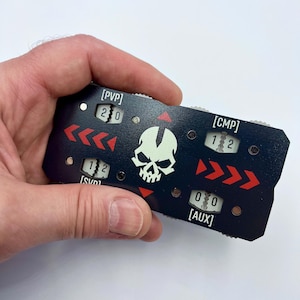 May include: A rectangular black gaming counter with a central white skull graphic. It features four mechanical dials labeled [PVP], [CMP], [SVD], and [AUX] with numerical displays. Red arrow icons point across the surface, and small metal screws secure the faceplate.