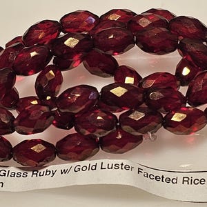 May include: Strand of faceted, dark red Czech glass beads with gold luster accents. The beads are rice-shaped, measuring 12x8mm. The beads are strung on a clear string.