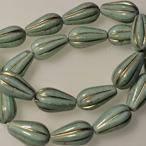 May include: Strand of teardrop-shaped beads in a pale teal color with gold stripes. The beads are strung together, creating a necklace or bracelet component. The beads have a glossy finish and are arranged in a curved pattern.