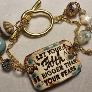May include: Gold-toned bracelet with a toggle clasp, featuring a rectangular ceramic charm inscribed with "LET YOUR FAITH BE BIGGER THAN YOUR FEARS." The bracelet includes various beads in blue, white, and brown, and star-shaped charms.