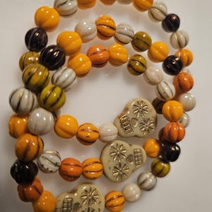 May include: A strand of beads featuring round, pumpkin-shaped pieces in shades of orange, white, black, and olive green, interspersed with three flat, cream-colored skull-shaped beads that have gold-colored floral and geometric detailing.
