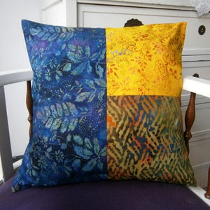 Peut inclure: Un coussin carré avec un motif patchwork. Le coussin est fait de quatre tissus différents, chacun avec un motif différent. Les tissus sont bleus, jaunes et bruns. Le coussin est posé sur une chaise blanche.