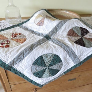 Può includere: Un quilt bianco con un bordo verde e quattro disegni circolari a patchwork. I disegni sono realizzati con tessuti di diversi colori e sono disposti a forma di girandola.