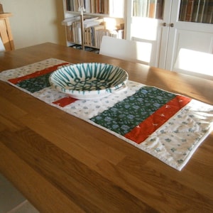 Peut inclure: Un chemin de table blanc avec des motifs floraux rouges, verts et bleus. Un bol en céramique blanc avec un motif vert et bleu est posé sur le chemin de table.