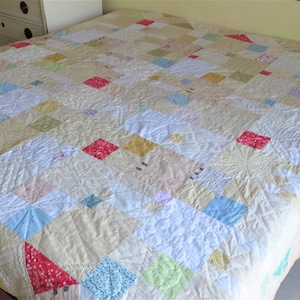 Op de afbeelding: Een handgemaakte quilt met een patchwork-ontwerp. De quilt heeft vierkanten in verschillende kleuren, waaronder rood, blauw, geel en groen, tegen een crèmekleurige achtergrond. De quilt ligt op een bed.