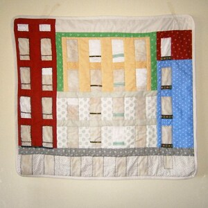 Peut inclure: Une courtepointe colorée avec un motif de bâtiments rouges, blancs et bleus avec des fenêtres. La courtepointe est faite de carrés de tissu cousus ensemble pour créer un motif de patchwork.