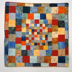 Op de afbeelding: Een kleurrijk patchwork-deken met een verscheidenheid aan vierkanten in tinten blauw, rood, oranje, geel en bruin. Het deken is gemaakt van veel verschillende stoffen met verschillende patronen.