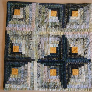 Peut inclure: Un coussin carré avec un motif de courtepointe en forme de cabane en rondins dans des tons de bleu, vert et blanc. La courtepointe a une bordure en tissu bleu foncé et présente un carré central avec un carré plus petit à l'intérieur. Le carré plus petit est rempli d'un tissu jaune clair.