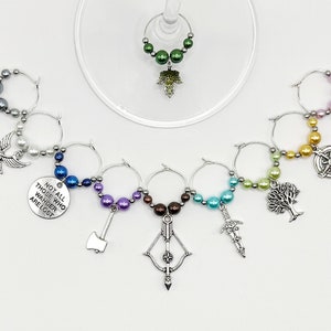 May include: A selection of wine glass charms featuring decorative beads and charms. Charms include a compass, tree, axe, bow and arrow, sword, and a disc with the text "NOT ALL THOSE WHO WANDER ARE LOST".
