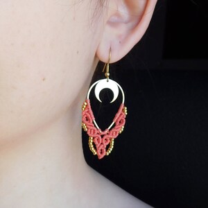 Op de afbeelding: Koraal en gouden oorbellen met een halve maan bedel. De oorbellen zijn gemaakt met macramé en hebben een delicate, bohemian stijl.