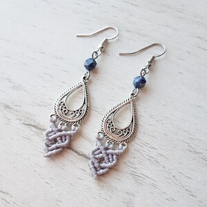 May include: A pair of silver earrings with a teardrop-shaped pendant and a blue gemstone bead. The pendant has a delicate filigree design and is accented with a gray macrame tassel.