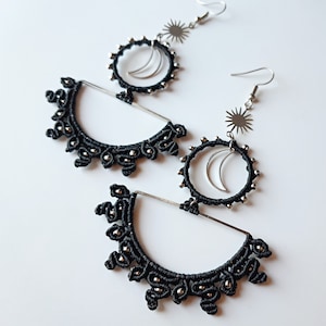 May include: A pair of black and silver earrings featuring a crescent moon and sun design. The earrings are made with black cord and silver beads.