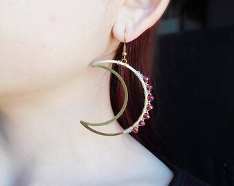 May include: Gold crescent moon earrings with red beads. The earrings are dangling from the earlobe.
