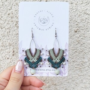 May include: A pair of handmade macrame earrings with green, brown, and white beads. The earrings are hanging from silver hooks and have a light green stone at the bottom. The earrings are on a white card with the text "Xanthi Elena Unique Jewelry" and "xanthielena.etsy.co".