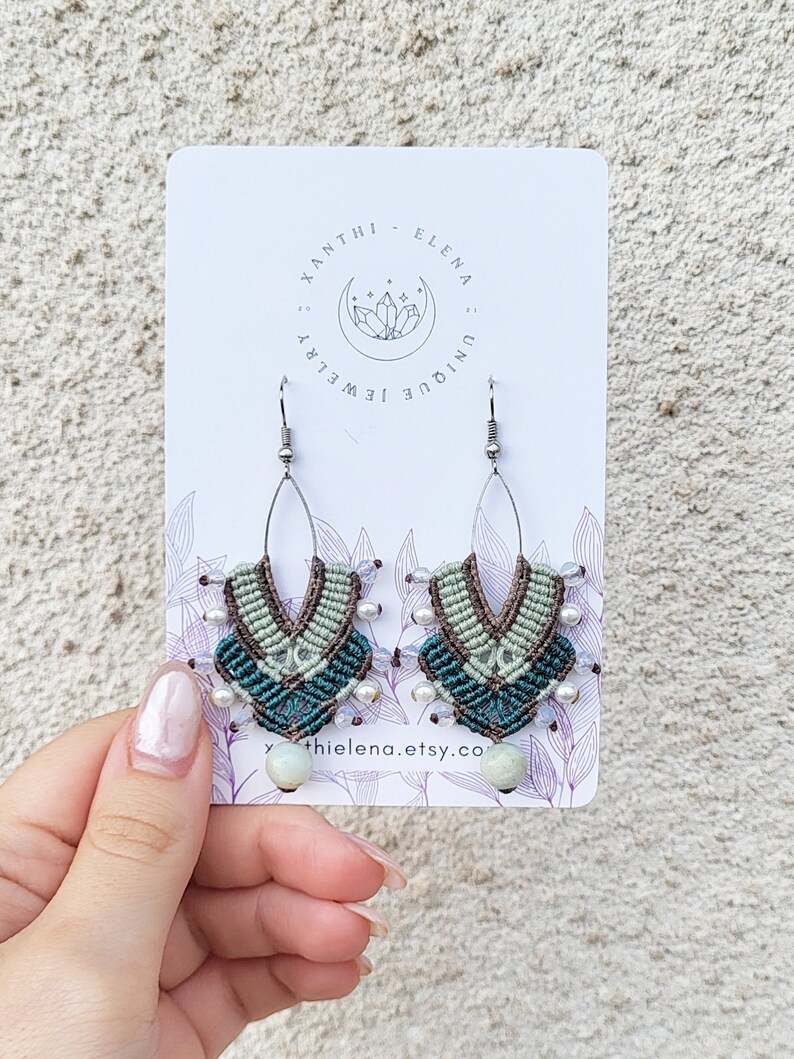 May include: A pair of handmade macrame earrings with green, brown, and white beads. The earrings are hanging from silver hooks and have a light green stone at the bottom. The earrings are on a white card with the text "Xanthi Elena Unique Jewelry" and "xanthielena.etsy.co".