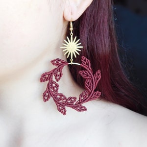 May include: Gold-colored sunburst earrings with a burgundy macrame leaf design. The earrings are hanging from a gold hook. The leaf design is a circular shape. The earrings are a unique and stylish accessory.