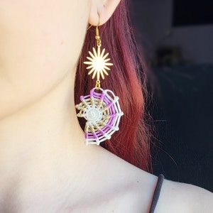 May include: Gold star charm dangle earrings with a purple and white macrame spiderweb design.