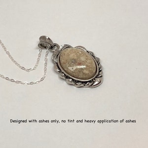 May include: A silver pendant necklace with a light brown oval-shaped stone set in a silver frame. The stone is made from ashes and has a textured surface.  "Designed with ashes only, no tint and heavy application of ashes" is written below the image.