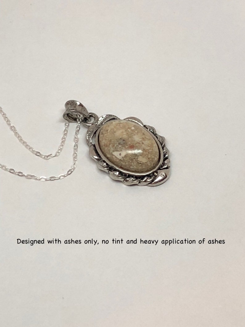 May include: A silver pendant necklace with a light brown oval-shaped stone set in a silver frame. The stone is made from ashes and has a textured surface.  "Designed with ashes only, no tint and heavy application of ashes" is written below the image.