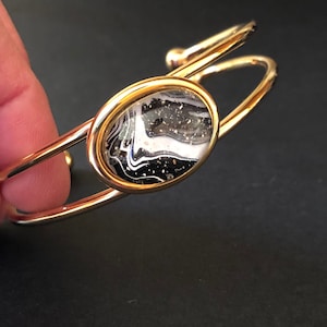 May include: A gold bangle bracelet with a round, black and white resin inlay. The inlay has a swirling pattern and gold flecks.