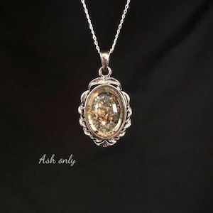 May include: A silver chain necklace with a silver oval pendant. The pendant has a clear glass insert filled with ash.