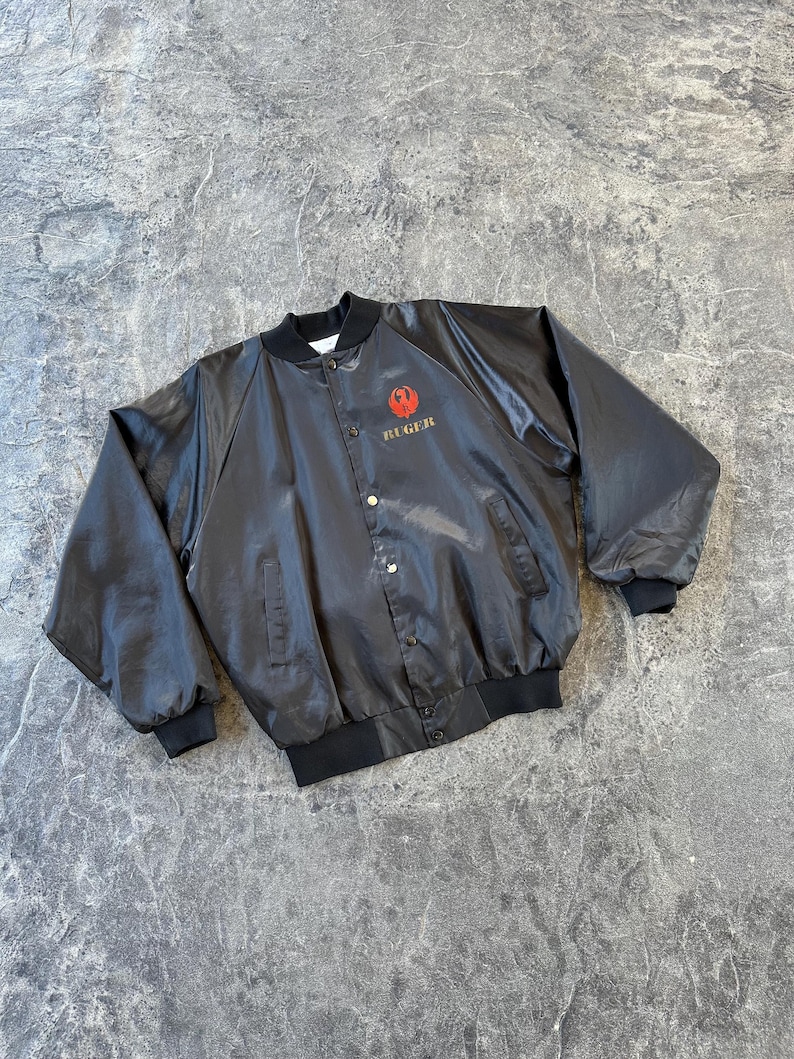 May include: A black satin bomber jacket with a red and gold Ruger logo on the left chest.