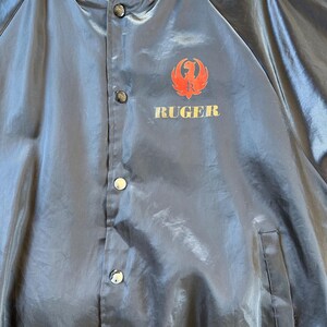 May include: A black satin bomber jacket with a red and gold Ruger logo on the front. The jacket has a button closure and a pocket on the left side.