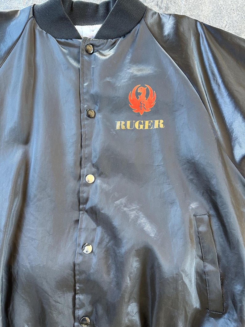 May include: A black satin bomber jacket with a red and gold Ruger logo on the front. The jacket has a button closure and a pocket on the left side.