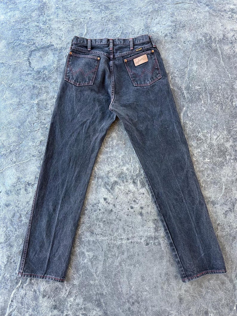 May include: A pair of dark wash denim jeans with red stitching detail and a leather patch with the Wrangler logo on the back.