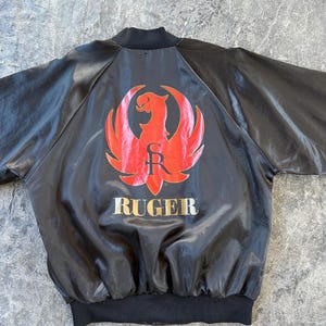 May include: Black satin bomber jacket with a red and gold Ruger logo on the back. The logo features a stylized eagle with wings spread and the word "Ruger" below it.