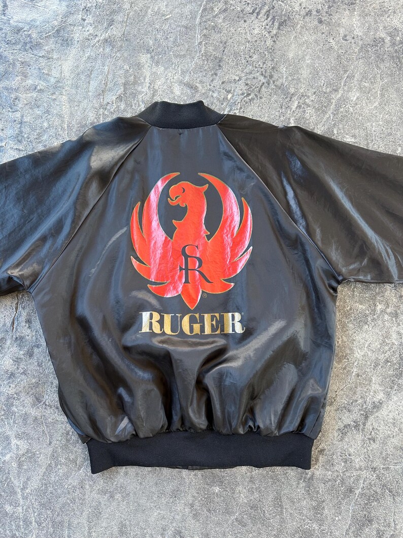 May include: Black satin bomber jacket with a red and gold Ruger logo on the back. The logo features a stylized eagle with wings spread and the word "Ruger" below it.