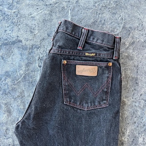 May include: Black denim jeans with red stitching and a brown leather back pocket patch with the Wrangler logo.