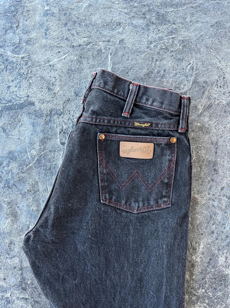May include: Black denim jeans with red stitching and a brown leather back pocket patch with the Wrangler logo.