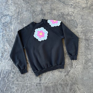 May include: Black sweatshirt with a blue, pink, and yellow flower design on the left shoulder.