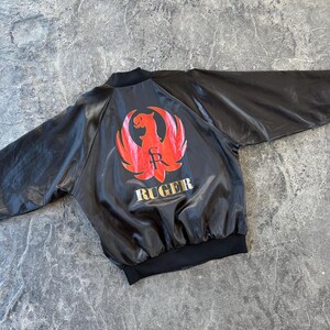 May include: A black satin bomber jacket with a red and gold Ruger logo on the back. The logo features a stylized eagle with the letters "R" and "S" inside the wings.