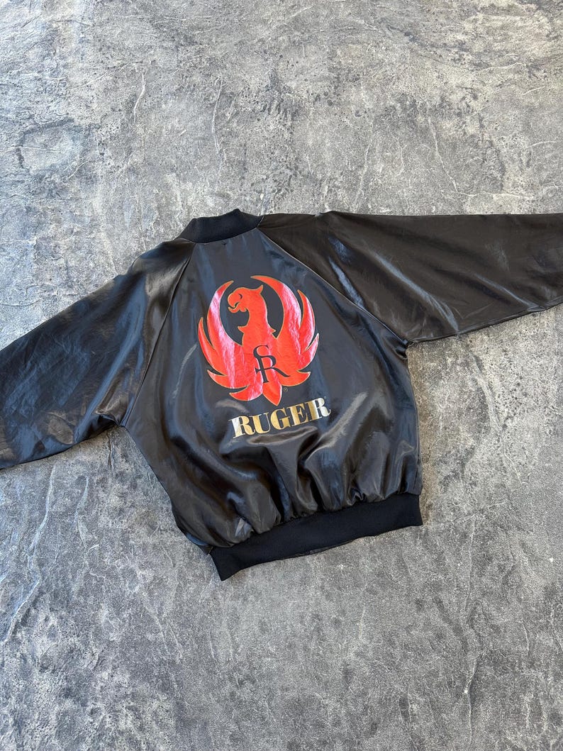May include: A black satin bomber jacket with a red and gold Ruger logo on the back. The logo features a stylized eagle with the letters "R" and "S" inside the wings.