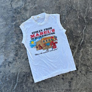 May include: A white sleeveless t-shirt featuring a graphic print with the text STOLEN FROM MABEL'S WHOREHOUSE. The design includes a building, a woman in a red dress, and the text RENO CHECK YOUR WEAPONS and Where the customer comes first.
