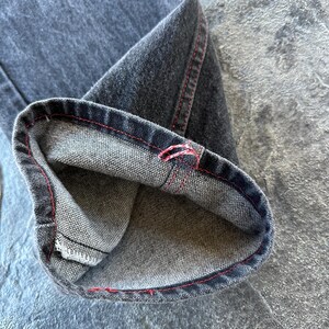 May include: Close-up of the inside of a pair of dark grey denim jeans with red stitching. The jeans are folded over to show the inside of the fabric.