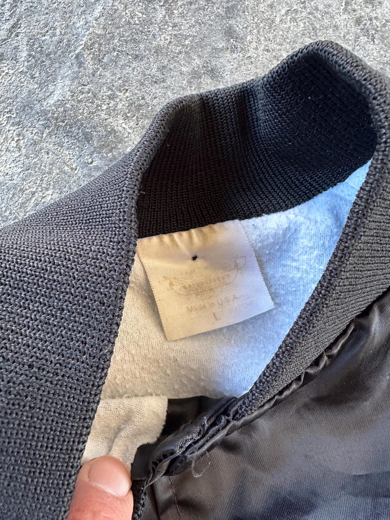 May include: A black satin bomber jacket with a ribbed collar and a white tag that reads "Manufactured in USA" and "L".