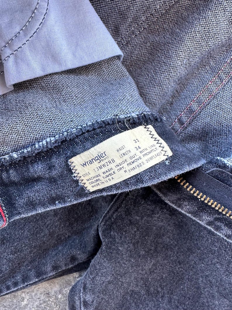 May include: A black pair of Wrangler jeans with a white tag that reads "Wrangler" and has the following information: "Style 13MWZRB, Waist 33, Length 34, Machine Wash Inside Out With Like Colours, Tumble Dry, Remove Promptly, Made in USA, R8HAF923 26WPL6428".