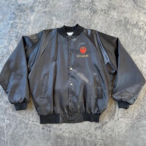 May include: A black satin bomber jacket with a red and gold embroidered Ruger logo on the left chest.