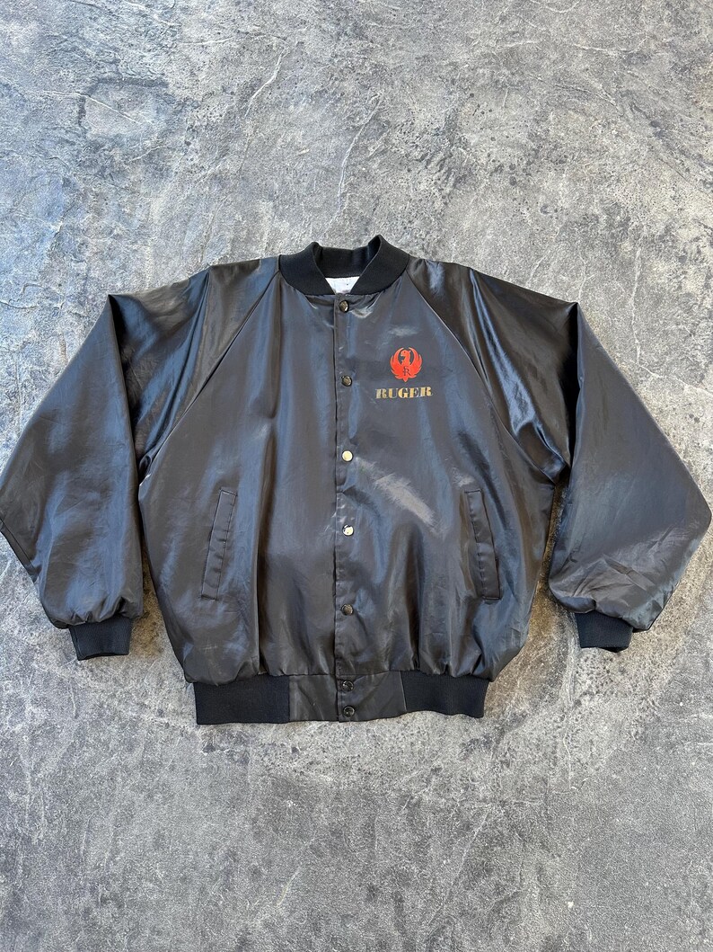 May include: A black satin bomber jacket with a red and gold embroidered Ruger logo on the left chest.