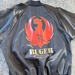 May include: Black satin bomber jacket with a red and gold Ruger logo on the back. The logo features a stylized eagle with the letter "R" inside a circle.