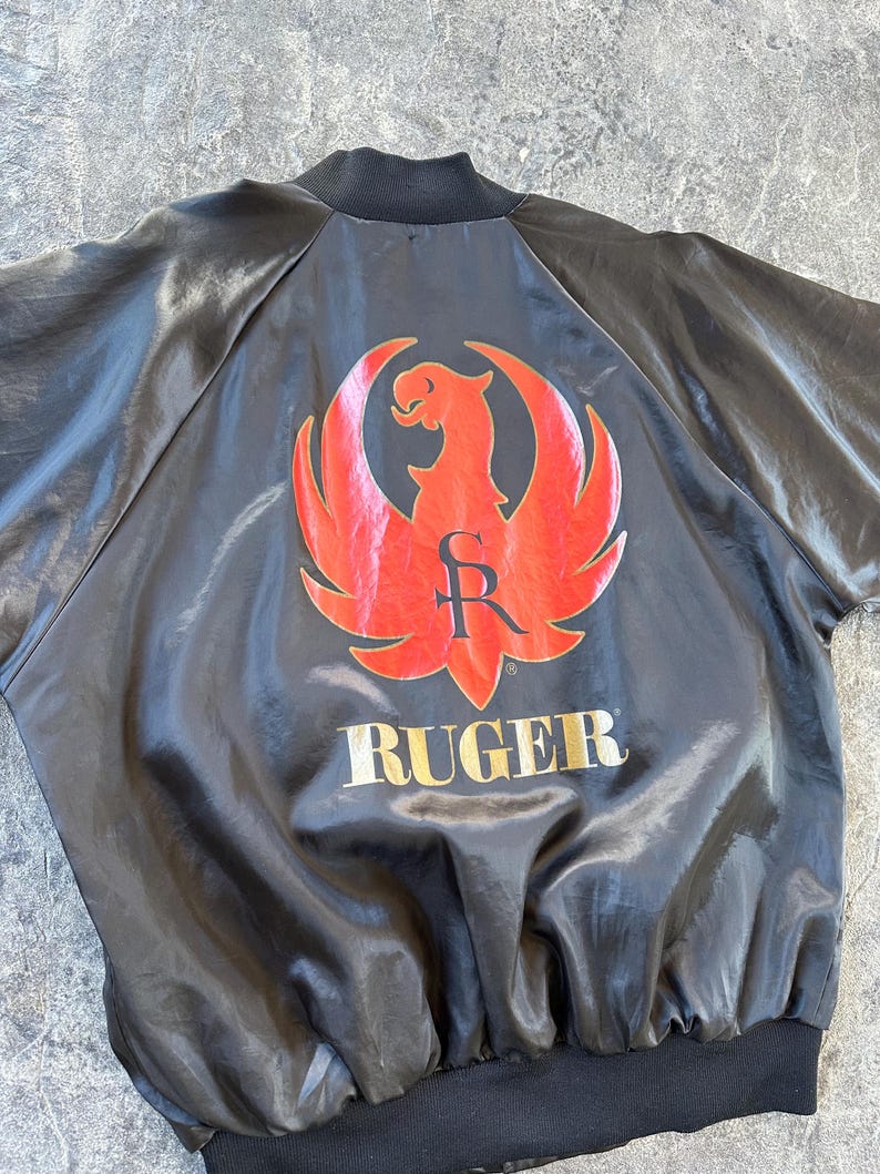 May include: Black satin bomber jacket with a red and gold Ruger logo on the back. The logo features a stylized eagle with the letter "R" inside a circle.