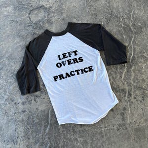 May include: A white baseball-style t-shirt with black three-quarter length raglan sleeves and a round neckline. The front of the shirt features the black text "LEFT OVERS PRACTICE" printed in a bold, sans-serif font across the centre chest area.